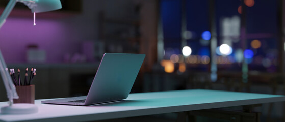 A laptop computer on a table in a modern dark office room at night features dim purple neon lights.