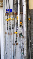 A Cluster Of Water And Electrical Meters Intertwined With Pipes On A City Wall. The Vertical Composition Highlights The Various Textures And Labels Under Bright Daylight