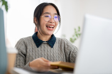 A cheerful young Asian female college student is studying online in a cafe co-working space.