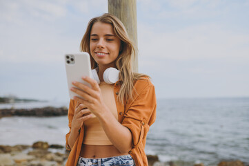 Young calm smiling woman she wears orange shirt casual clothes listen to music in headphones use mobile cell phone walk on sea ocean sand shore beach outdoor seaside in summer day. Lifestyle concept.