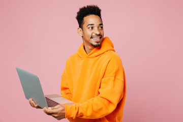 Side view young IT man of African American ethnicity wear yellow hoody casual clothes hold use work on laptop pc computer look aside isolated on plain pastel pink background studio. Lifestyle concept
