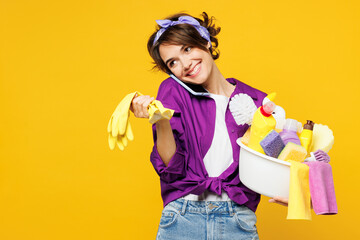 Young smiling woman wears purple shirt hold basin with detergent bottles do housework tidy up talk...