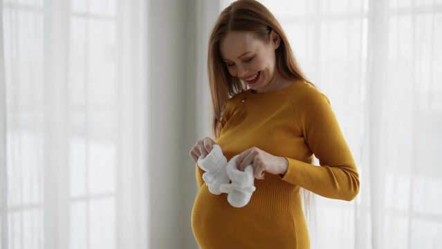 Pregnant woman enjoying expecting baby. Happy female future mother holding child knitted soft white booties in hands with smile, tenderly stroking tummy. Happy pregnancy, expecting baby concept.