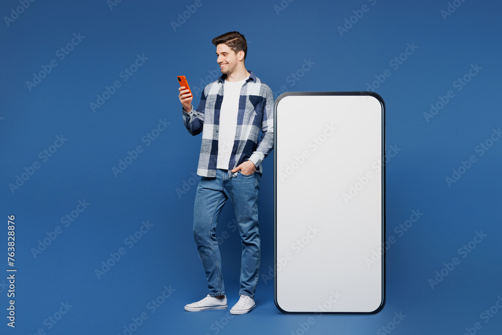 Canvas Prints Full body young happy man he wear shirt white t-shirt casual clothes stand near big huge blank screen mobile cell phone with area using smartphone isolated on plain blue background. Lifestyle concept.