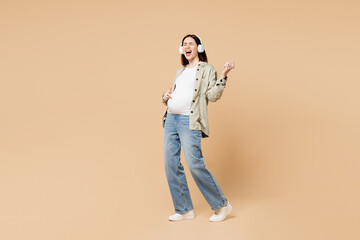 Fototapeta premium Full body young pregnant woman future mom wear grey shirt headphones with stomach tummy with baby listen music play air guitar isolated on plain beige background. Maternity family pregnancy concept.