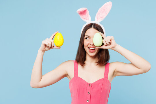 Young surprised smiling woman she wear pink casual clothes rabbit bunny ears hold in hand look at painted eggs isolated on plain blue cyan background studio portrait. Lifestyle Happy Easter concept.