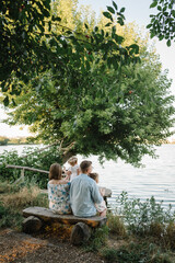 A happy family is sitting back on a bench on the shore of a lake and looking at a beautiful sunset view. Mom, dad hug children daughter near pond. Mother, father embrace kids and spend time together.