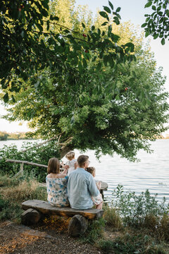 A happy family is sitting back on a bench on the shore of a lake and looking at a beautiful sunset view. Mom, dad hug children daughter near pond. Mother, father embrace kids and spend time together.