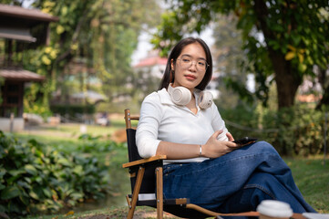 An Asian woman relaxing in her backyard, sitting on a camping chair with a smartphone in her hand.