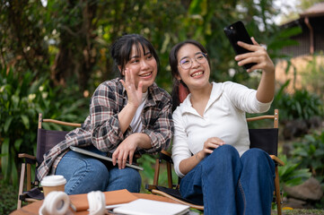 Two cheerful Asian female friends enjoy taking selfies with a smartphone together outdoors.
