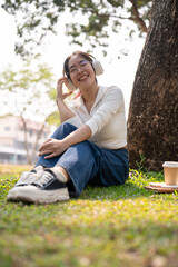 A carefree Asian woman is enjoying the music on her headphones, sitting on the grass under a tree.