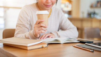 A cropped shot of an Asian woman sipping coffee while reading a book at a table in a coffee shop.