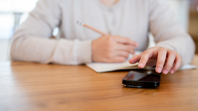 A close-up image of a woman using her phone and writing in a book at a wooden table in a cosy room.