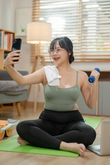 Fototapeta na wymiar Beautiful Asian woman in sportswear is taking selfies with her smartphone while exercising at home.