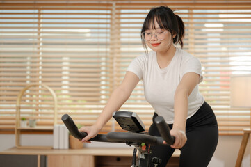 Fototapeta na wymiar A strong Asian woman in sportswear is exercising on a fitness bike, doing indoor cardio at home.