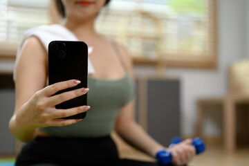 Select focus on hand of sports woman using mobile phone while lifting dumbbell in living room