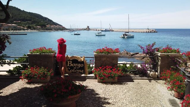 Elegant tourist woman in red with hat, Marciana Marina village, the flowery district Borgo al Cotone: meaning COTTON VILLAGE. Tourist on travel in Italy. Marciana Marina town in summer on Elba island