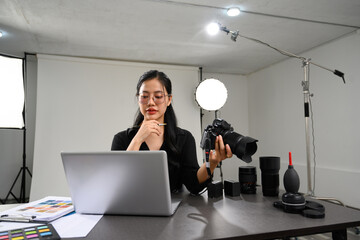 Creative young woman holding her DSLR camera and working with laptop at photo studio interior
