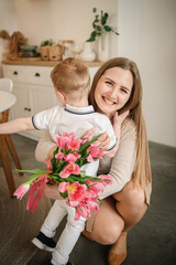 Son hugging smiling mom. Happy family holiday. Cute little child boy greeting mother in kitchen. Beautiful kid gives mother a big bouquet of tulip flowers and makes surprise for Mother's Day.