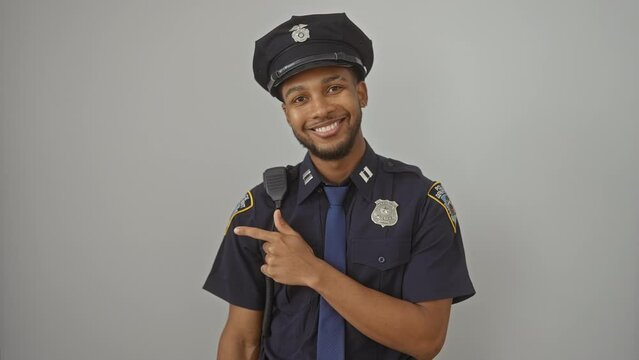 Cheerful african american man all suited up in police uniform, flashing a big smile and pointing finger to the side, on an isolated white background