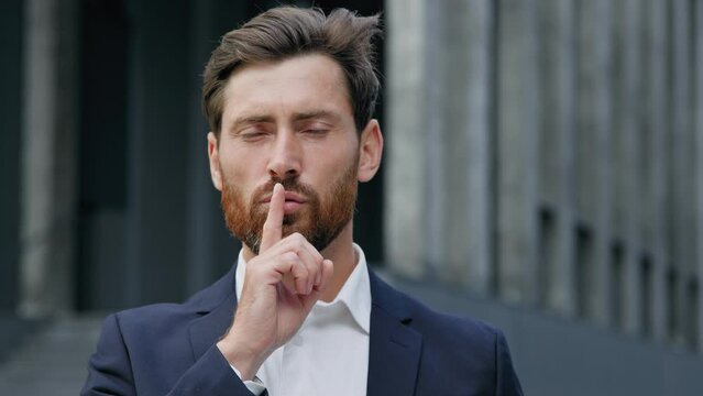Mysterious Caucasian man businessman guy leader boss showing silence sign holding finger to mouth make gesture hush Shh forbid to speak business secret confidential information outdoors male portrait