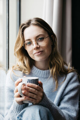 Portrait of a young beautiful blonde woman holding a cup of tea by a window inside their home, in an intimate and cozy atmosphere.