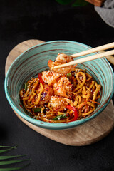 Chinese noodles with shrimps and vegetables. Person holding chopsticks with shrimp over asian fried udon. Asian food with holding prawns. Close-up shrimp on fried noodles