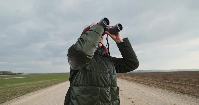 female ornithologist birdman watches birds with binoculars on dirt gravel road against a background of a stormy sky