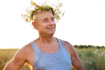 happy cool satisfied trendy funky hipster mature man of 60 years old smiles in T-shirt, cute wreath wildflowers, daisies, natural crown on head, image faun, satyr in sunlight, connection with nature - obrazy, fototapety, plakaty