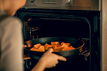young woman in an apron puts a dish with pumpkin in an electric oven for baking in kitchen