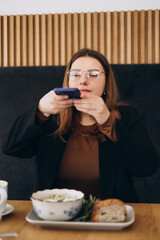 Female blogger influencer hold phone take food mobile photo on phone sit at cafe table. Woman...