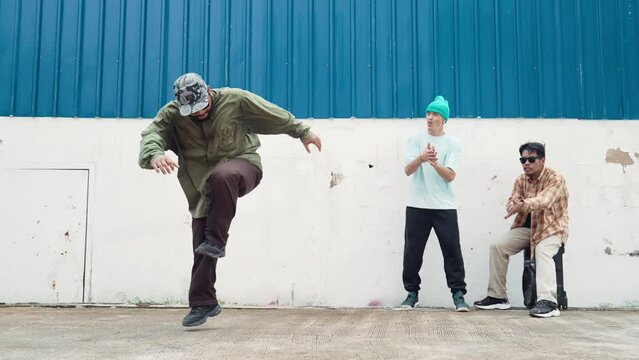 Skilled hipster doing cart wheel while multicultural friends encourage him. Hip-hop dancer moving foot step to music surrounded by group of hipster at wall near street. Outdoor sport 2024. hiphop.