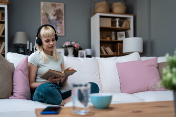Young woman relaxing on a sofa at home. Listening music on wireless headphones and reading book....