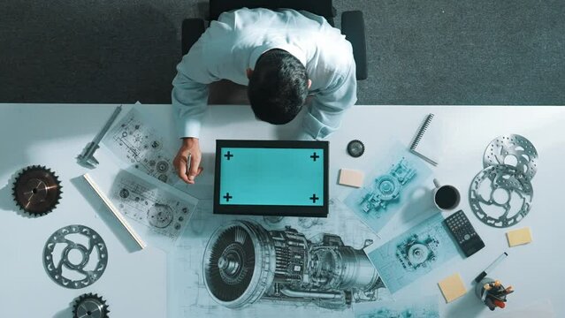 Top view of skilled engineer writing in paper while looking at tablet display green screen with turbine engine sketch design and metal gear and architectural equipment scatter around. Alimentation.
