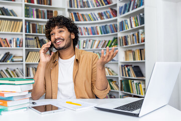 Happy young man with curly hair makes a phone call while working on his laptop in a home office with bookshelves in the background.