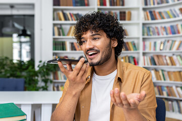 Cheerful young man with curly hair speaks into his smartphone using voice command in a library...