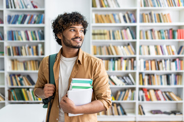 Young male student with curly hair smiling and holding textbooks in a modern library setting, exuding confidence and preparedness.