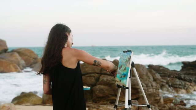 Female artist paints seascape on canvas at rocky shore. Waves crash, woman in casual dress enjoys painting hobby, captures ocean beauty. Art creation, landscape inspiration in serene nature. Slowmo