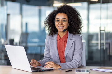 Friendly professional woman with curly hair, wearing a headset and working on a laptop in a modern office setting, looking at camera.