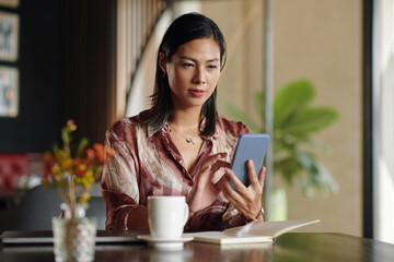 Businesswoman sitting at cafe table, checking emails on smartphone and writing in planner