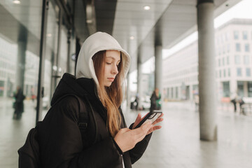 Half length portrait of girl in casual wear holding smartphone for blogging in the city in the autumn-winter season. Teen surfing the internet outdoors.