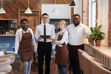 Medium long group portrait of young multi-ethnic waiting staff posing for camera at work in modern...