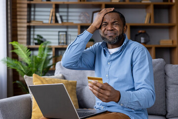 Adult male at home looking at his laptop with a concerned expression, holding a credit card,...
