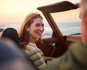 Retired Senior Couple On Vacation Sitting In Classic Sports Car At Beach Watching Sunrise