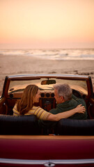 Rear View Of Retired Senior Couple On Vacation In Classic Sports Car At Beach Watching Sunrise