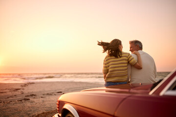 Rear View Of Retired Senior Couple Sitting On Hood Of Classic Sports Car At Beach Watching Sunrise