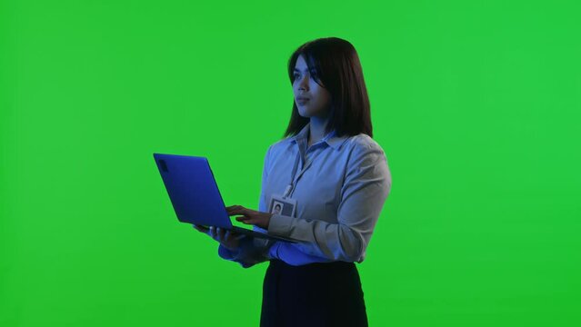 Female data center engineer wearing badge and formalwear typing on laptop while standing under blue neon light against green chroma key screen