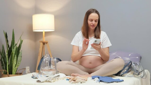 Adorable pregnant woman packing for hospital and taking notes in smart phone getting ready for newborn birth preparing clothes necessities holding cellphone in hands