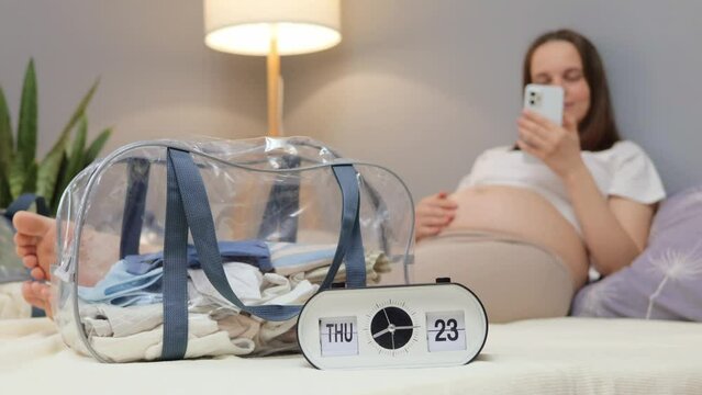 Closeup of clock with young pregnant woman with smart phone in hand lying at bed with baby clothes in bag on background reading information about childbirth in internet