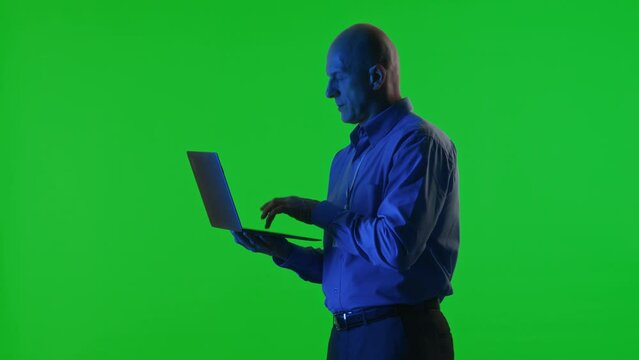 Data center specialist standing under blue neon light, holding laptop and typing code in studio with green chroma key background